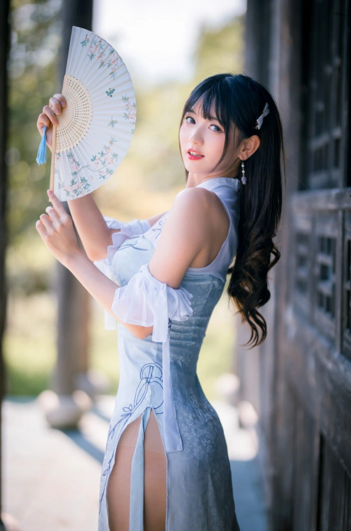 kyokyo身穿浅蓝色改良旗袍站在木质长廊旁,手持折扇,侧身回望镜头,阳光从侧后方照射