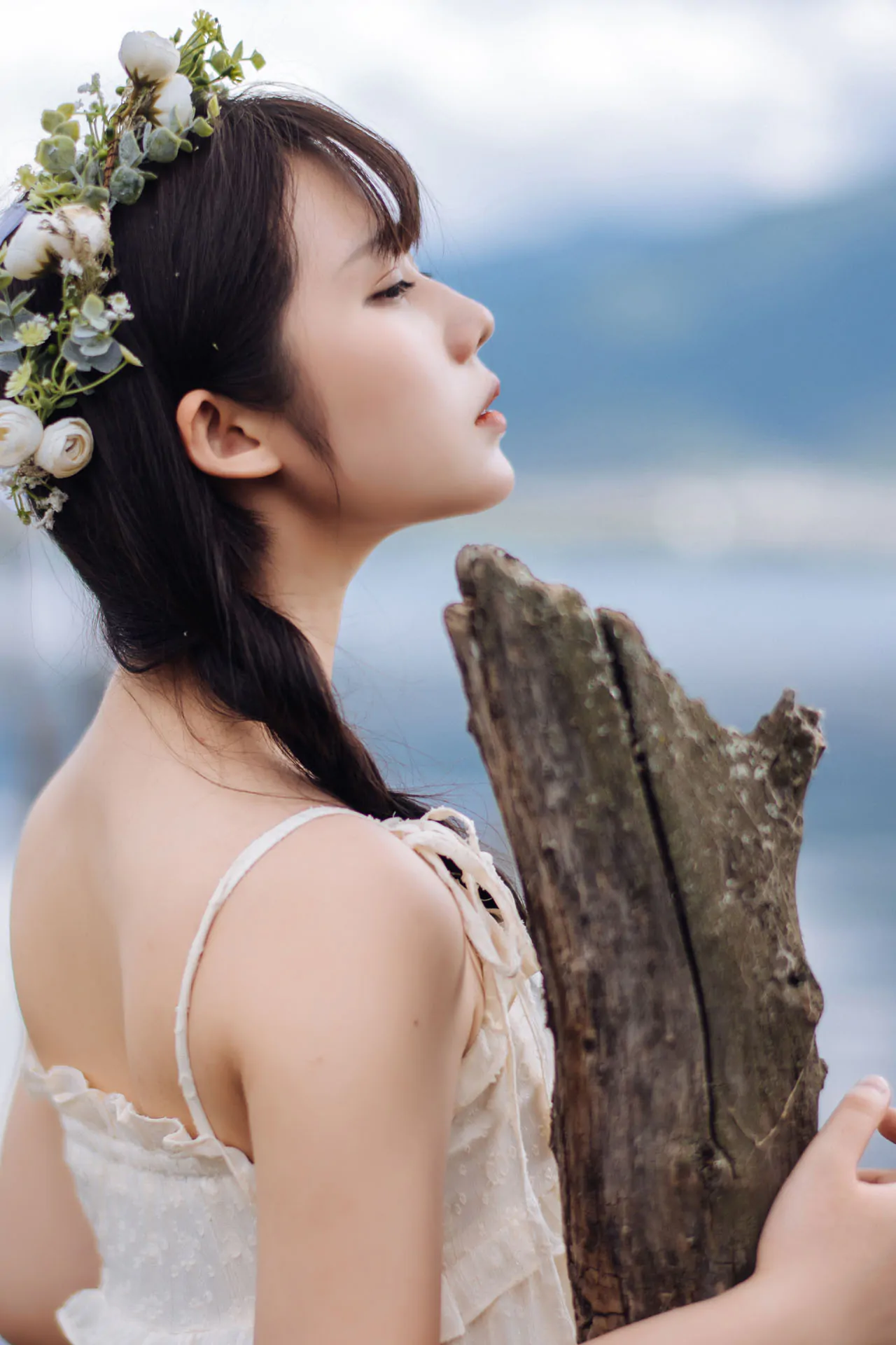 白色吊带连衣裙，头戴花环，侧脸靠近枯木，背景为远处山水虚化