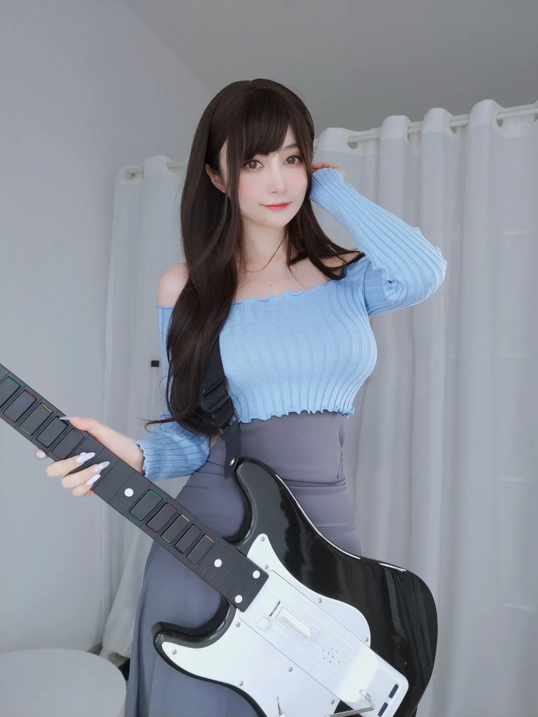 白银81蓝色露肩针织上衣与灰色高腰裙造型，手持黑色游戏吉他控制器，室内白色窗帘背景
