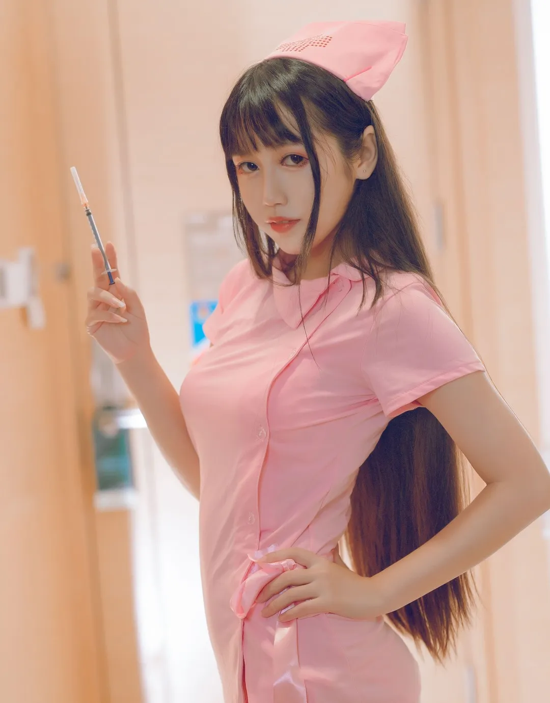 不呆猫粉色护士服 戴粉色护士帽 手持注射器 侧身站立 长直发 背景为暖色走廊与门框虚化