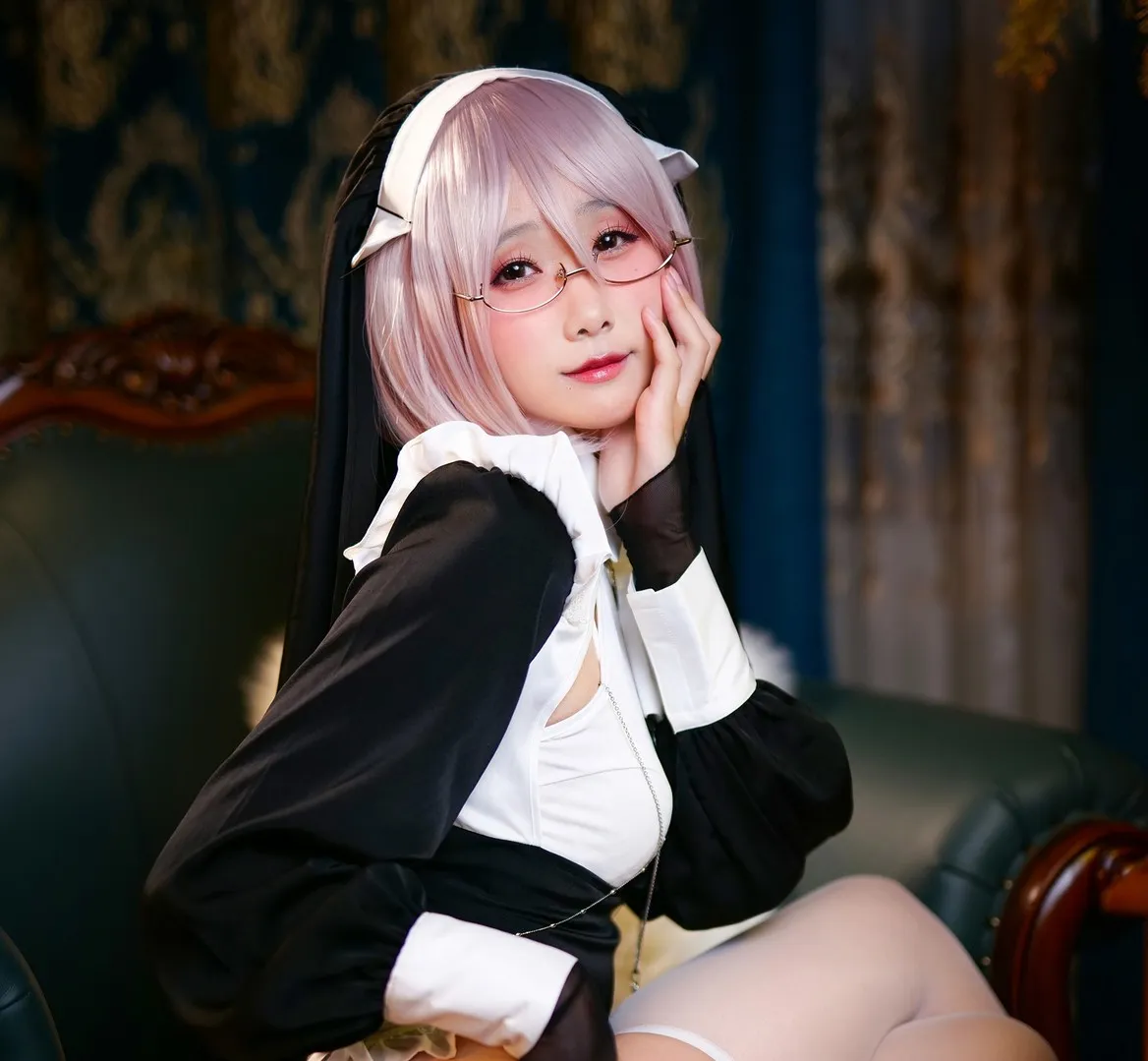 曉美媽粉色短髮造型佩戴金屬細框眼鏡，穿黑白修女風角色服裝坐在深色沙發上，手托臉頰，背景為厚重窗簾