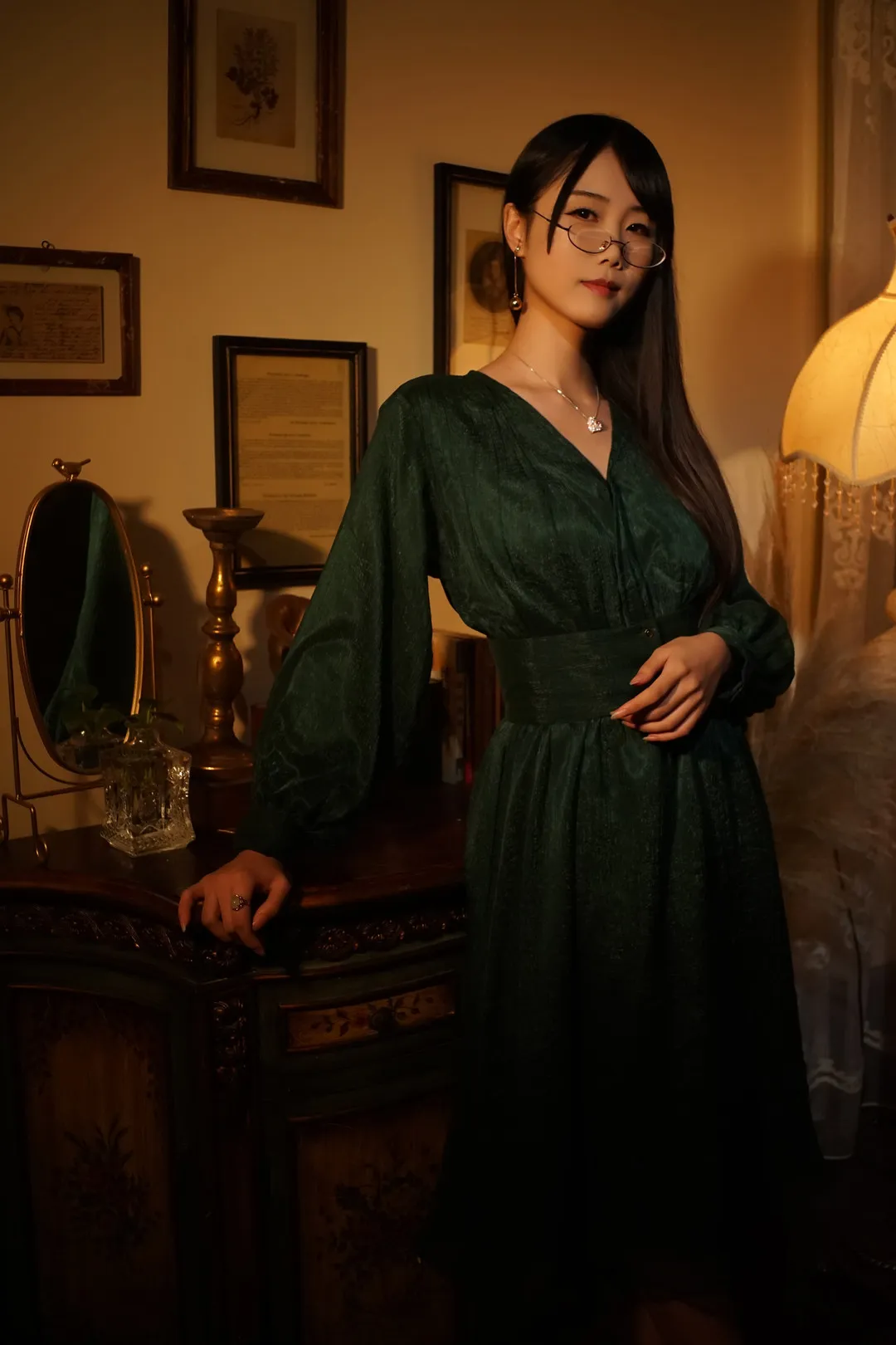 曉美媽身穿墨綠色長袖收腰連衣裙站在木質櫃旁,暖色檯燈側光照射,佩戴圓框眼鏡與項鍊,背景為復古相框與書櫃