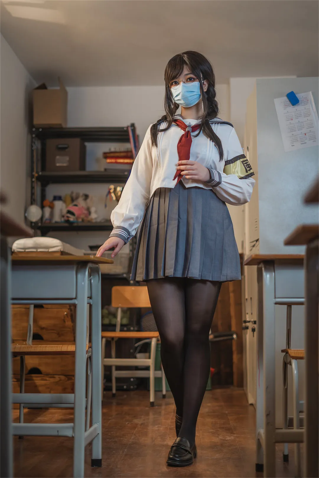 啊日日_Ganlory穿白色水手服上衣灰色百褶裙黑丝皮鞋站在教室过道佩戴口罩与眼镜手持红色领巾背景有课桌与置物架暖色顶灯