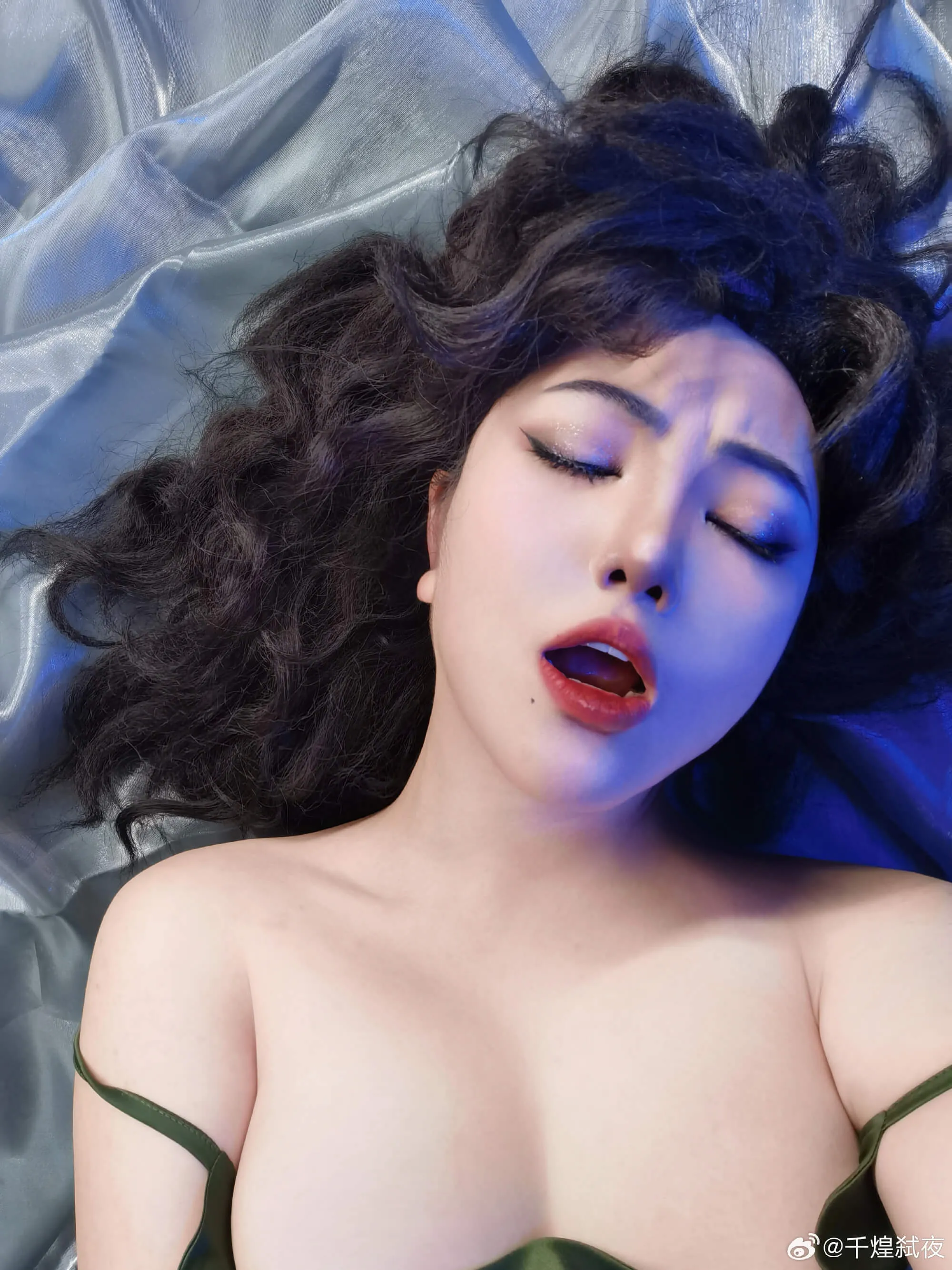 千煌弑夜躺在灰色缎面布料上，蓝光从侧面扫过，闭眼张口的戏剧表情，红唇与亮片眼妆对比强烈