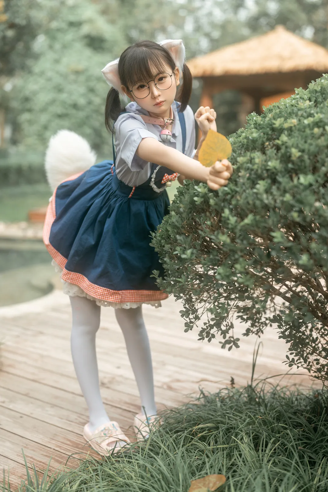 幼水铃衣在院子里戴猫耳眼镜穿蓝色裙装伸手拨树叶的照片