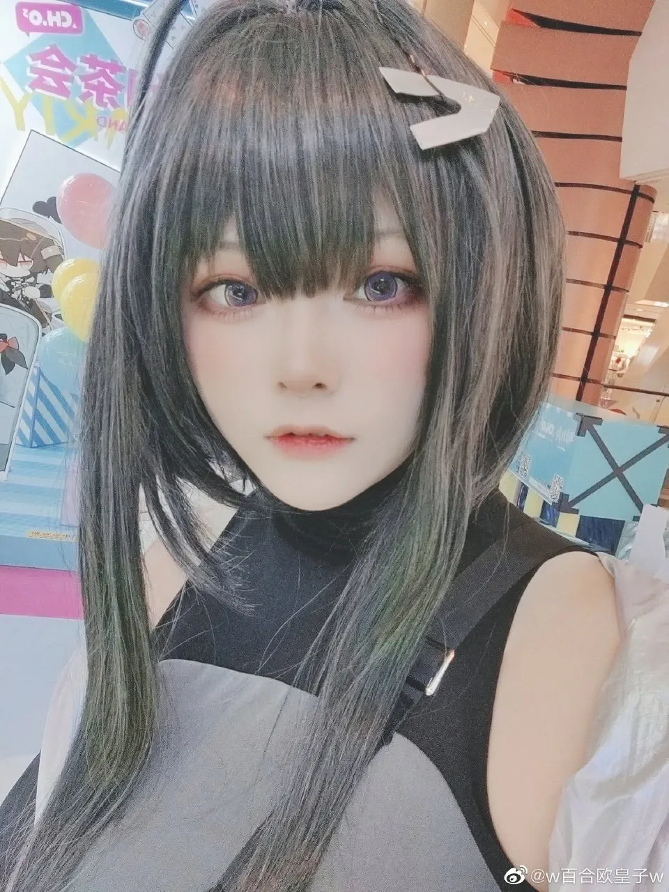 w百合欧皇子w在展会背景前自拍出镜，深色长发配黑灰无袖高领服装
