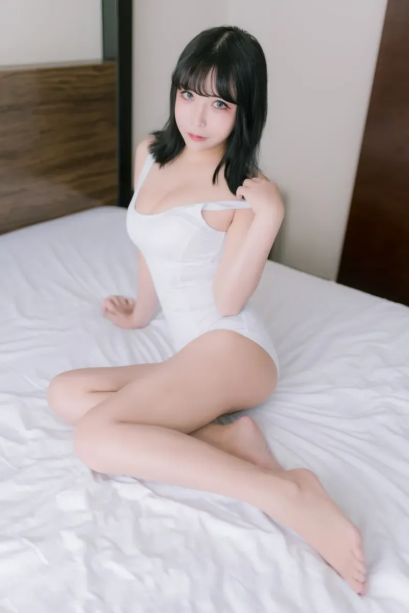 KuukoW 居家白色连体服写真图，人物留黑色中长发，穿白色连体服坐在白色床铺上，背景有木质墙面和深色门板。