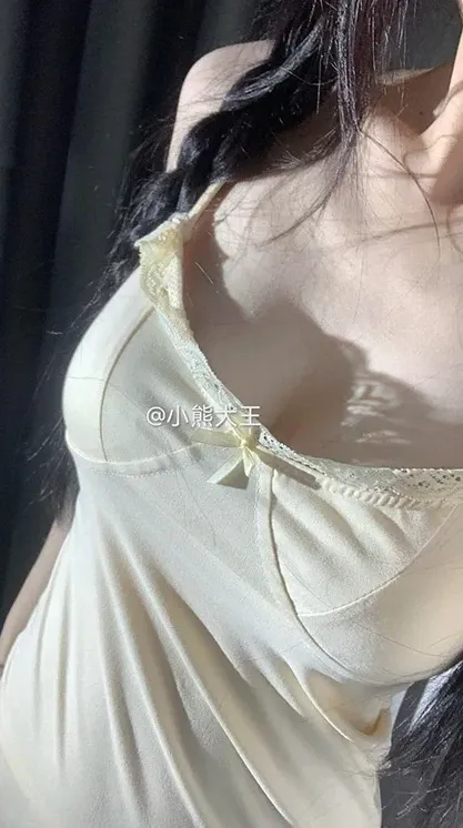 抖音小熊大王黑色长发穿浅黄色吊带服装,站在深色背景前侧身拍照